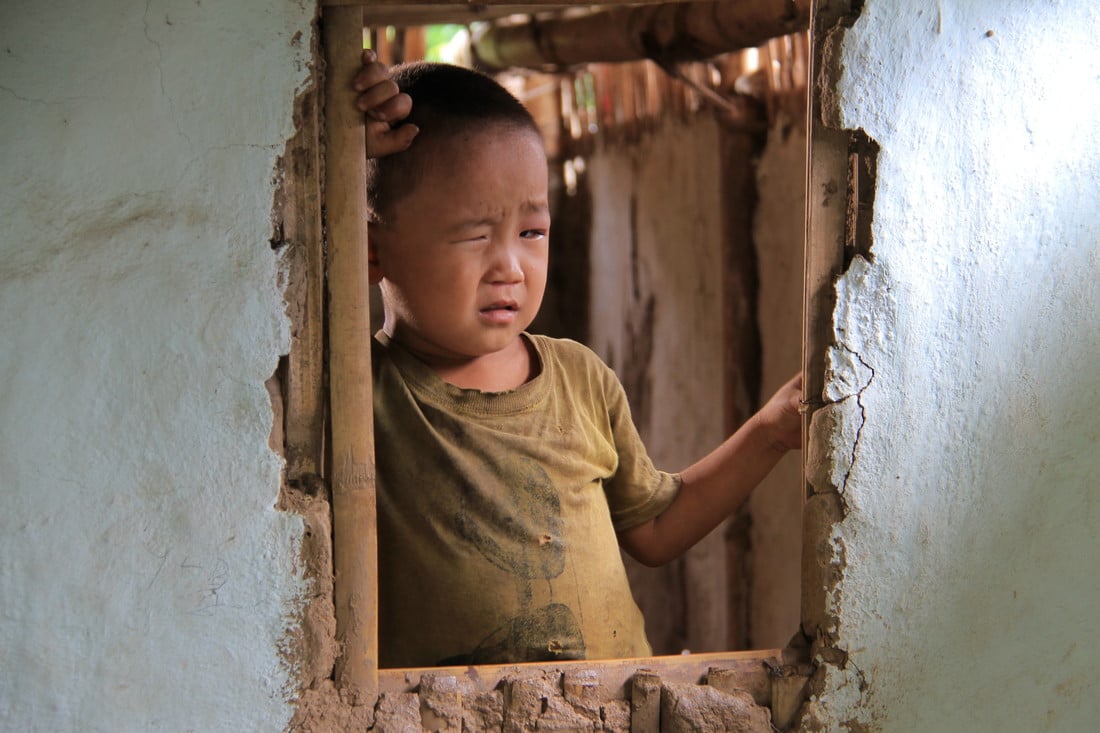 Sudip standing holding the sides of a window without glass in it, squinting.