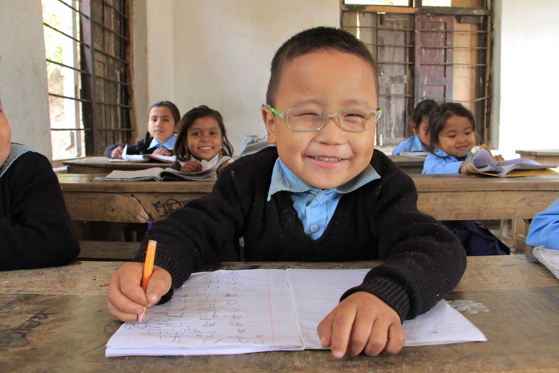Sudip sat at a desk in class, smiling and wearing glasses.
