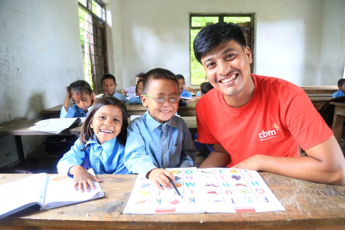 Sudip sat at his school desk with a CBM community worker and fellow student, all smiling.