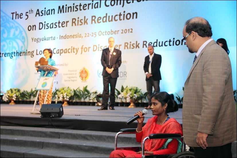 Kazol is wearing a red dress and sitting in a wheelchair. She is sat beside a stage, speaking into a microphone.