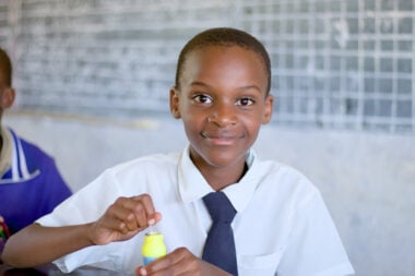 A little girl in school uniform smiles. She has a bubble bottle in her hands.