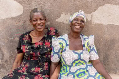 Two women sit down in front of a wall with the arms interlinked - both are smiling.