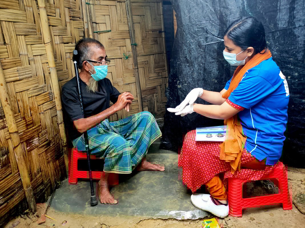 Man holding a walking stick sitting down and being shown how to wash hands effectively by a woman wearing PPE
