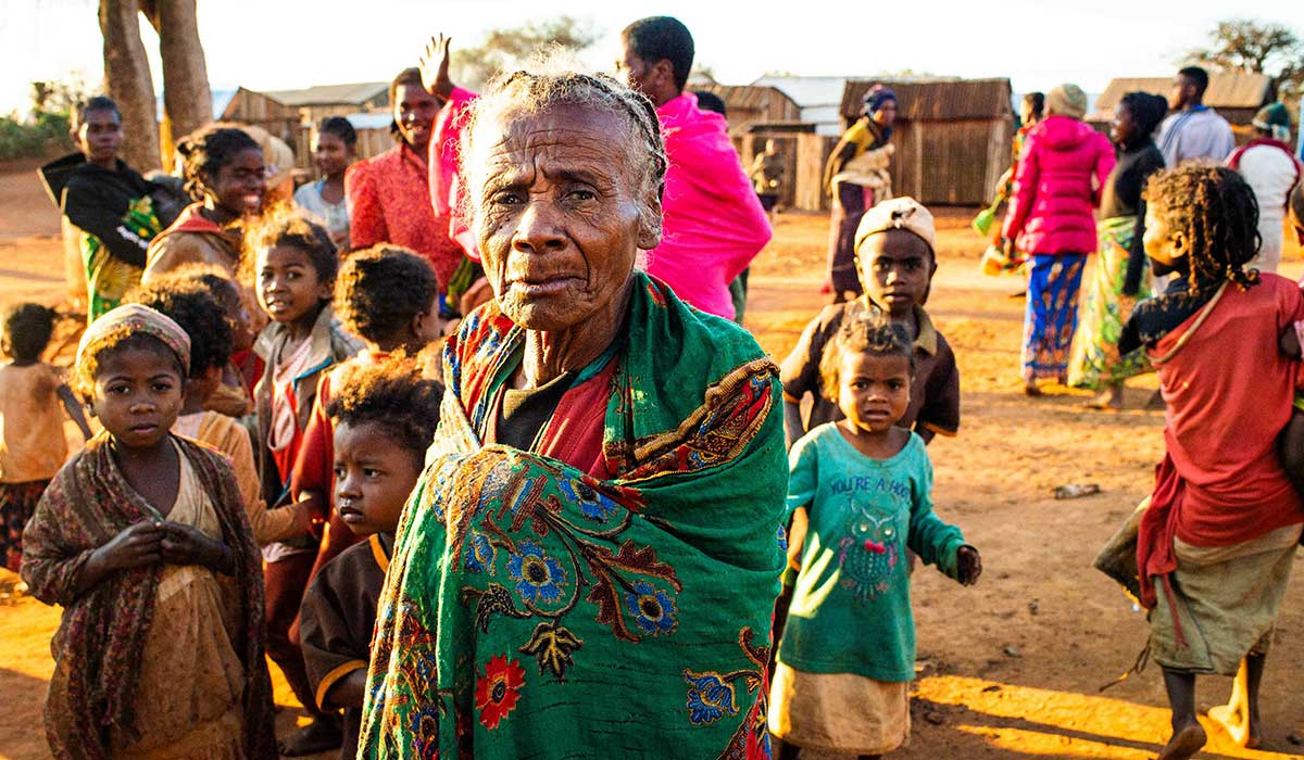 Elderly lady looks straight at the camera and stands in front of a group of children.