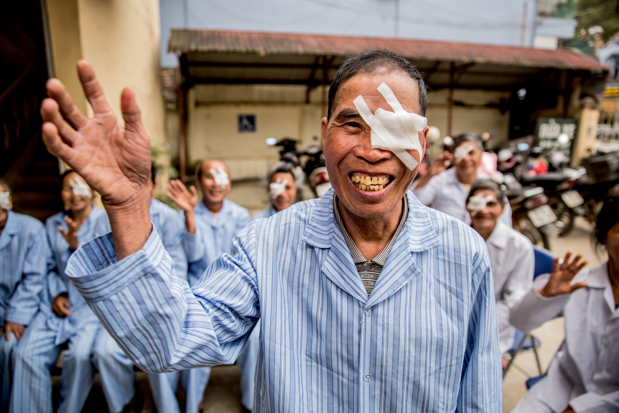 Khut smiling and waving, in a blue hospital gown with an eye patch over his left eye