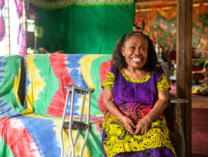 A woman from fiji wearing a purple and yellow dress with her crutches. She is is sitting down on a colourful sofa.