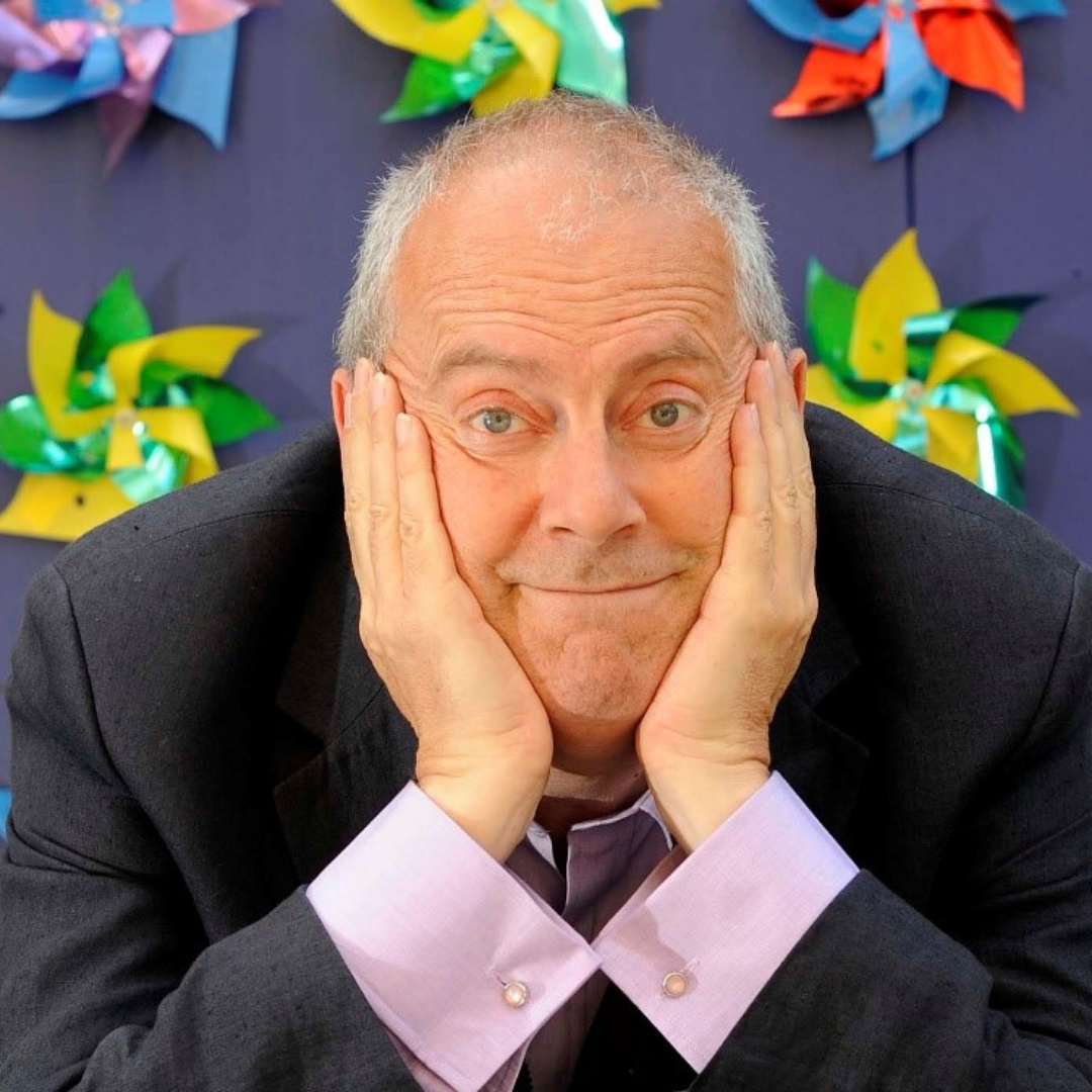 Broadcaster Gyles Brandreth leaning on his hands with a purple background.