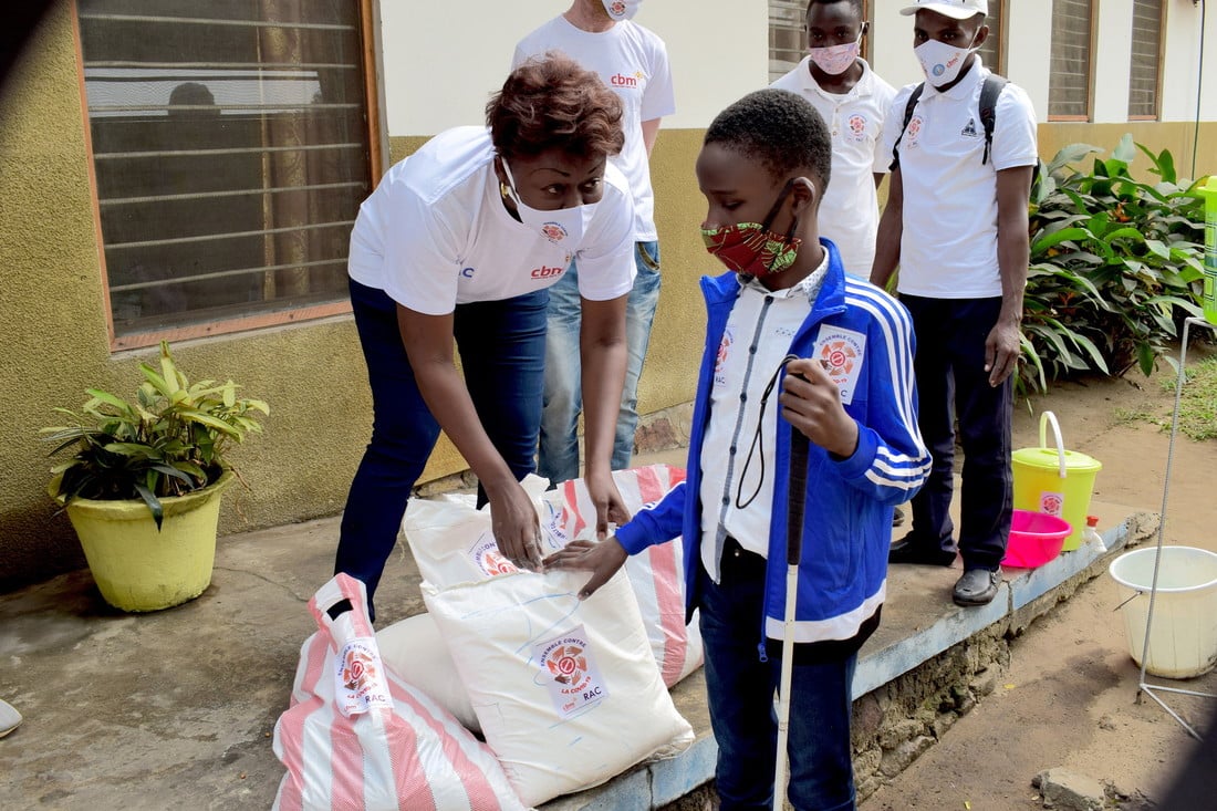 Woman wearing a face mask and CBM t-shirt handing food parcels to a boy with a white cane