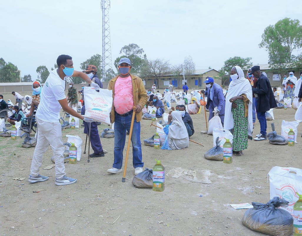 A man wearing  face mask hands a man with a walking stick emergency aid parcels, in a field in Ethiopia with lots of other community members standing nearby