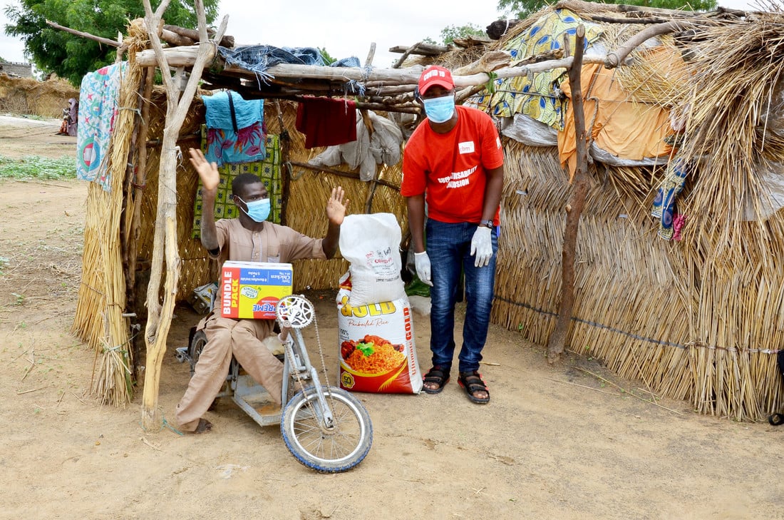 Male humanitarian worker delivering food parcels to a man with a physical disability sat on a tricycle in front of his home in rural Nigeria