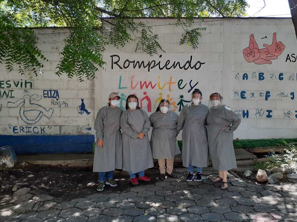 Five female volunteers standing in matching grey dresses and face masks, in front of a sign saying Rompiendo Limites.