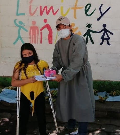A young girl on crutches being given emergency relief items by a man wearing PPE 