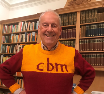 Gyles Brandreth wears a red and yellow jumper with 'CBM' on the front. He has his hands on his hips and smiles at the camera.