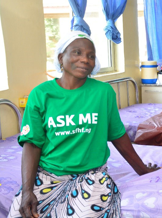 Justina wearing a green t-shirt which says 'Ask Me' on the front. She is sitting on a hospital bed, smiling.