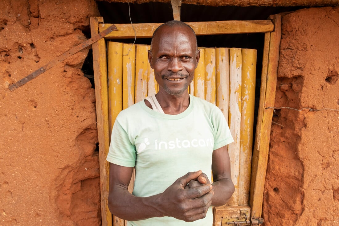 Julius holding his hands together and standing outside a wooden door attached to his mud hut.