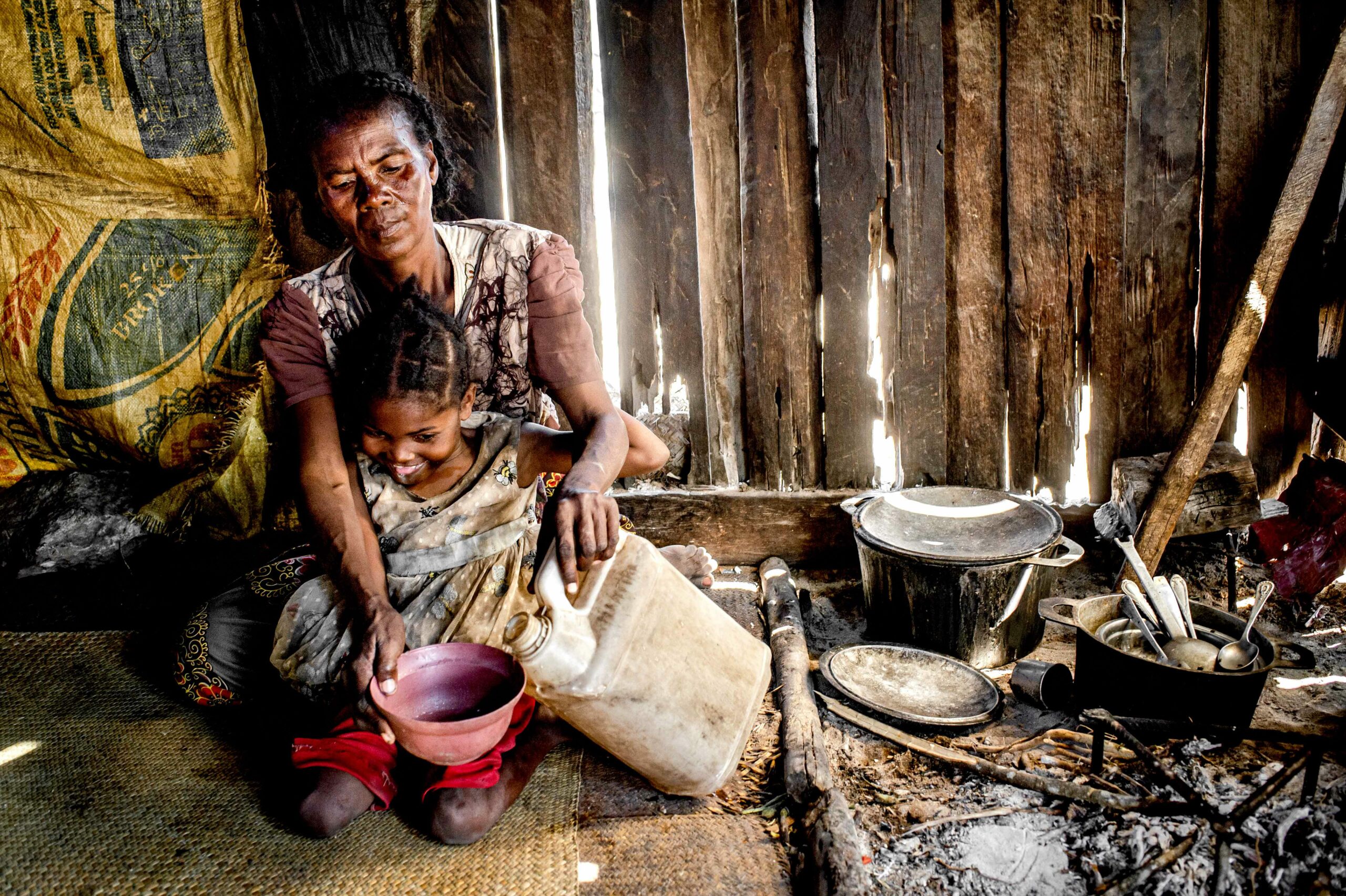 A women sits in her wooden hut, with her child on her knee, pouring something into a bowl