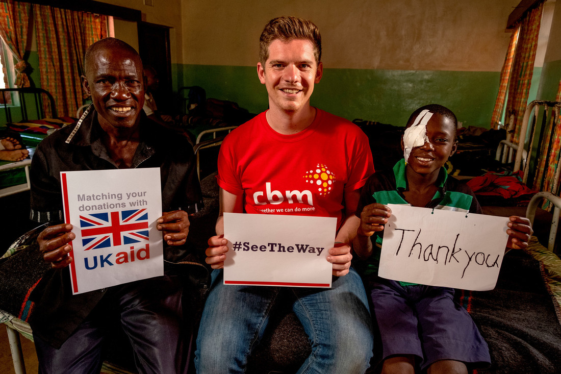 L-R: Jabes' father holding a UK Aid Match logo sign, CBM staff member holding #SeeTheWay sign and Jabes holding a Thank You sign.