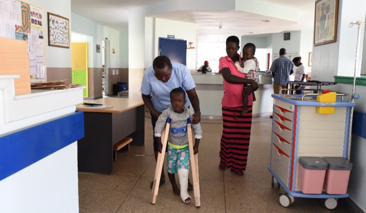 In the hospital, Nurse Florence has his arms around Makoma’s shoulders, guiding him to use crutches that rest under his arms, his leg in a plaster cast. 