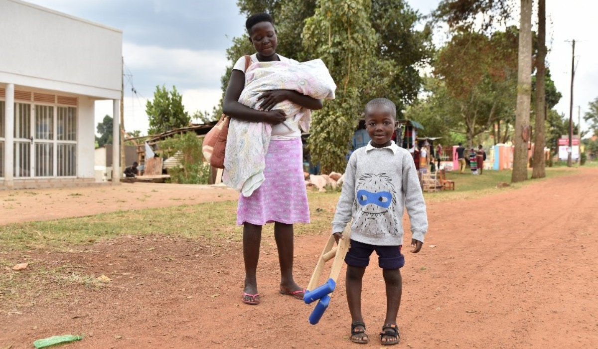Makoma smiles, standing straight without using the crutches he holds at his side, outside the hospital building with his mother who holds her baby in blankets