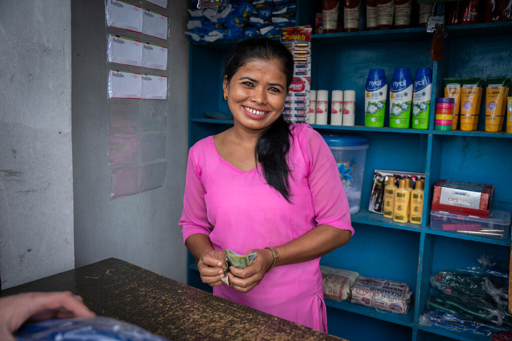 Chandrakala, wearing a pink shirt and holding money, stands in front of some goods and smiles at the camera