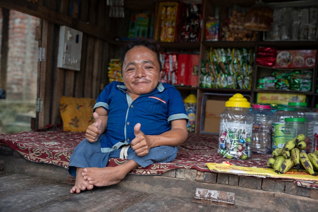 Deepak is wearing a blue shirt and is sitting on a rug next to jars of sweets and a bunch of bananas
