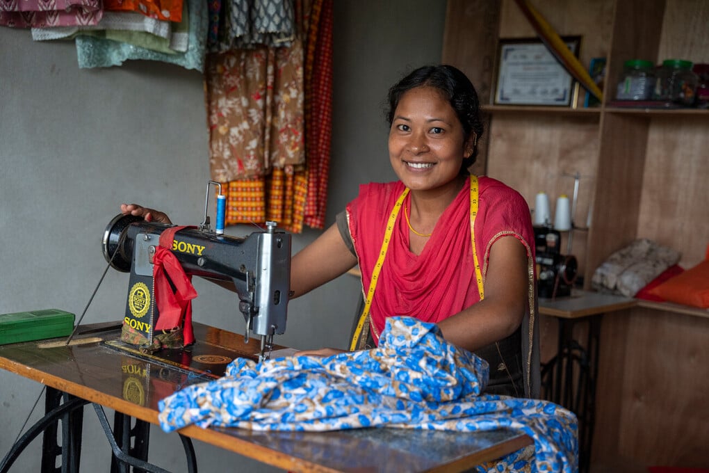 Lalita is wearing red and is using a sewing machine to alter a blue garment