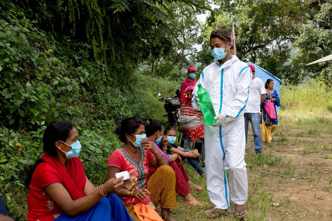 A male volunteer wearing PPE walks along a line of people sat on the floor, giving them water whilst they wait to be seen.