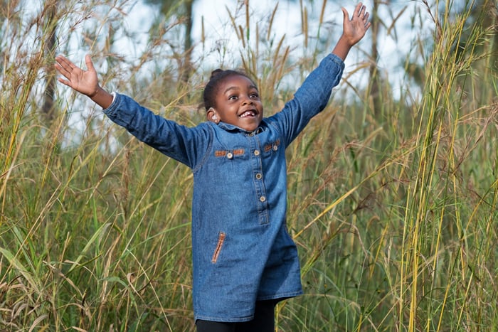 Six year old Tobile is outdoors surrounded by long grass, her arms thrown in the air, a radiant smile on her face