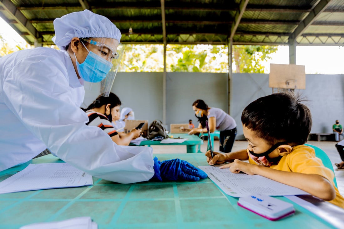 Female health worker wearing PPE sitting across a desk from a young boy wearing a face mask and filling out a form