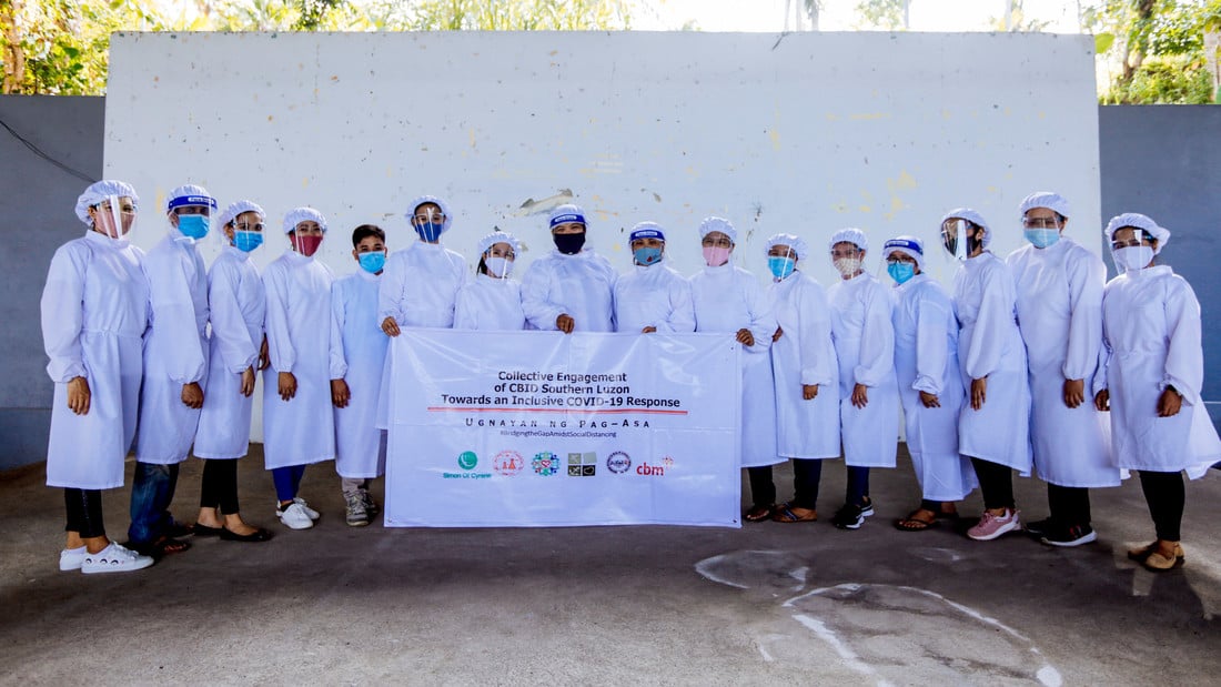 Group of 16 health workers wearing PPE and posing in front of a sign