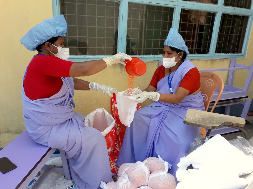 Two women wearing PPE, sat measuring out food into individual emergency relief parcels. 