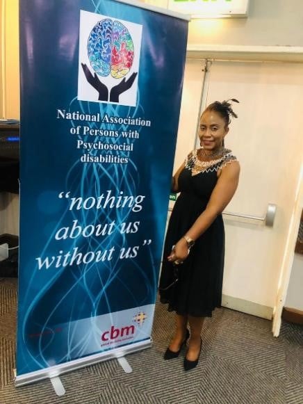 Tarisai Bere standing in front of a banner which says National Association of Persons with Psychosocial disabilities "nothing about us without us"