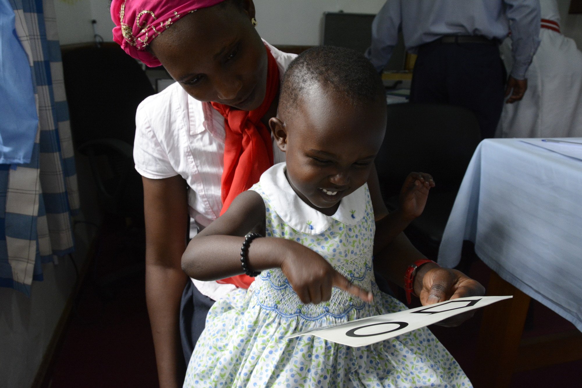 A young girl sat on her mum's lap, reading an eye chart
