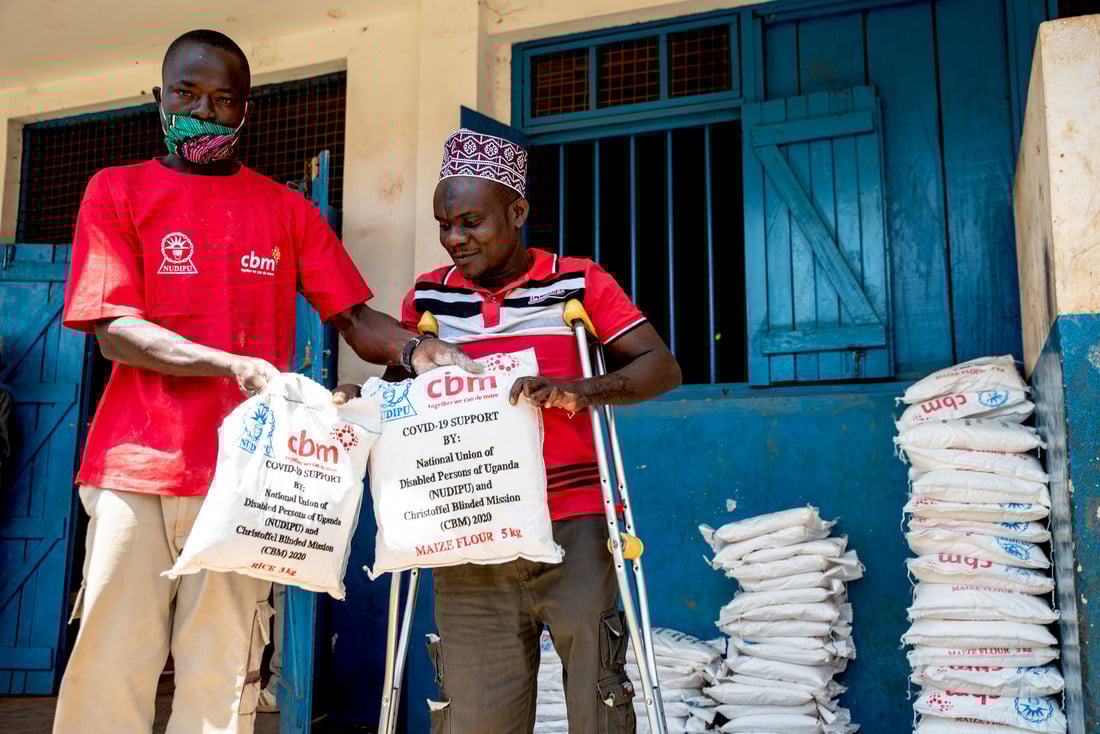 Man in a red CBM t-shirt hands a man using crutches 2 food parcels.