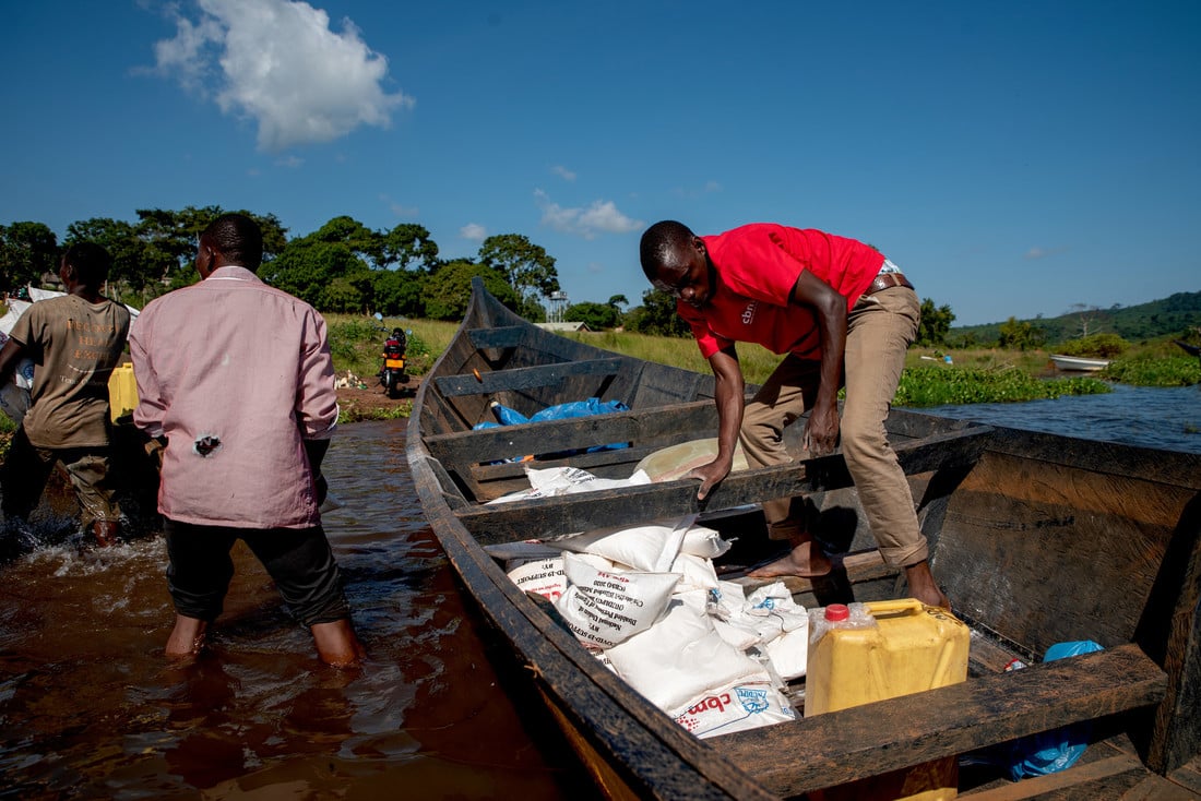 Man in a red t-shirt offloads relief parcels from a small boat on shore.