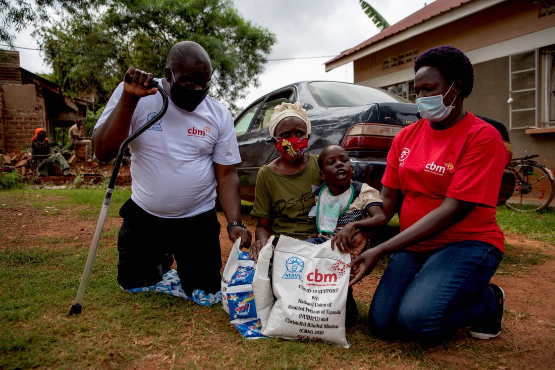 L-R: Male humanitarian worker wearing a white CBM t-shirt and holding a walking stick, elderly grandmother, 8-year-old Arthur on his grandmother's lap and female humanitarian worker wearing a red CBM t-shirt. All sat on the floor in front of emergency relief parcels.