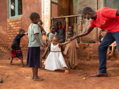 A girl wearing a white dress is in front of a house. Around her are other children and a man in a cbm t shirt who is holding her hand. Her other hand is held by another child who is also having his hand held to make a chain.