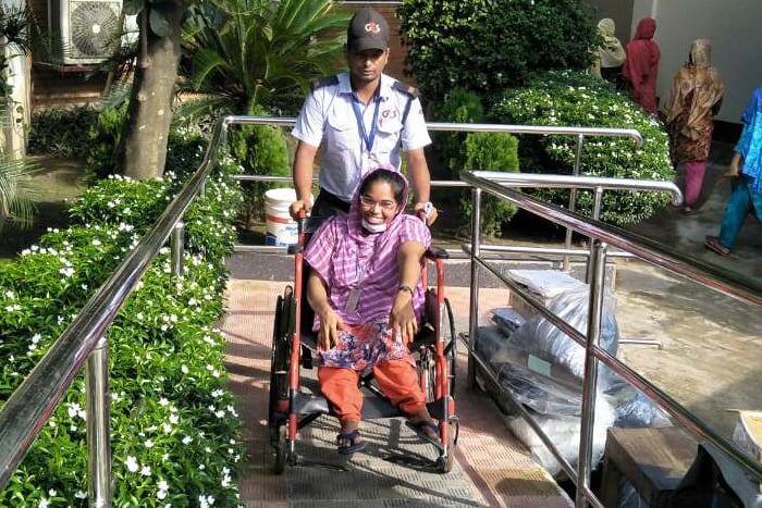 Accessible entrance, Bangladesh. A woman in a wheelchair is pushed along a ramp. She smiles broadly