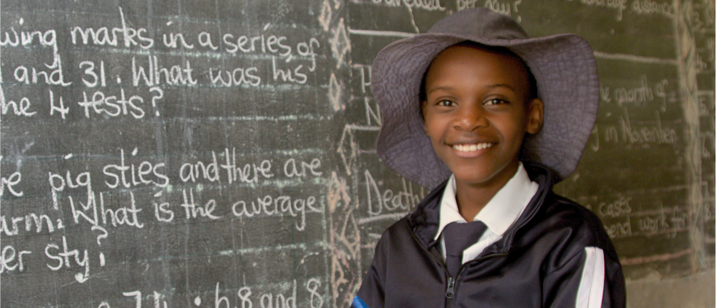 A girl stands in front of a chalkboard in a hat and school uniform.