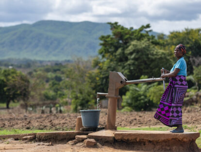 A woman in colourful clothing uses a manual hand pump to collect water. In the background are hills.