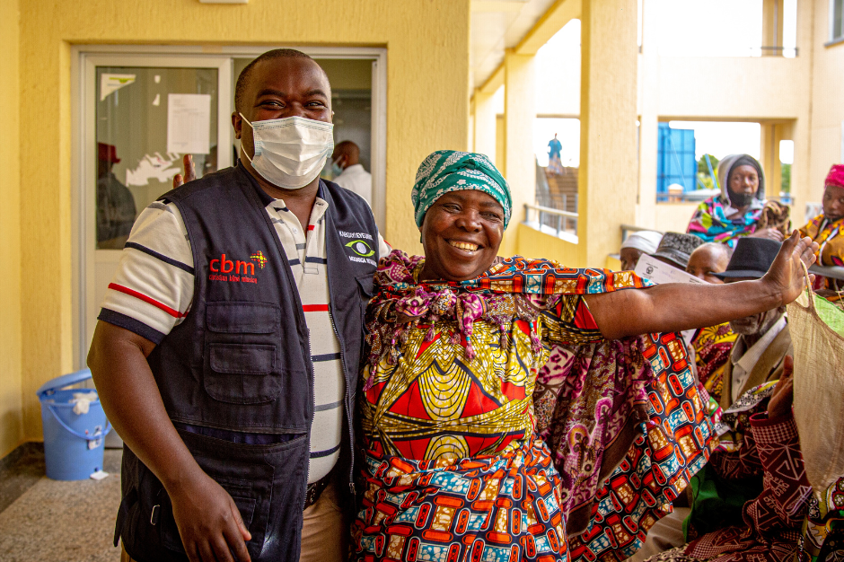 A doctor in a surgical mask and a woman in brightly coloured clothes smile and spread their arms wide.
