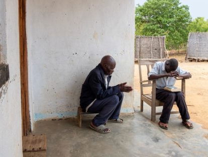 Cataract patient Batwel prays with his son Lemani. Batwel is sat on a footstool and has his hands together. Lamani is sat on a chair with his head down and a book in his lap.