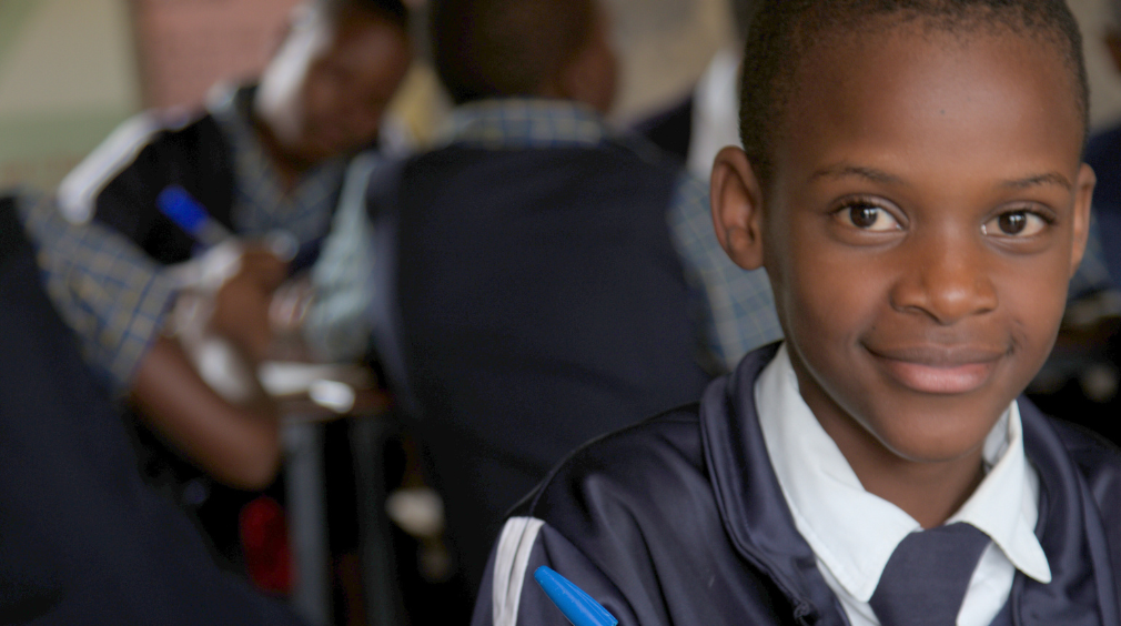 Tinotenda sits at a table with a pen in her hand. There are other students sat in the background.