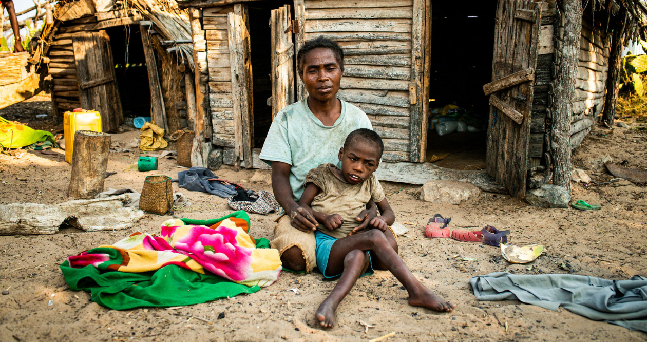 A mother sits with her child in her lap on the ground. Some household items are around them in front of a wooden house.