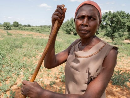 A lady stands in a field, looking into the camera while holding onto a wooden stick.