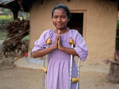 A young girl from Nepal, holding crutches underneath her arms. She is praying with her hands.