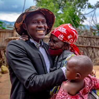 Velena holds her young granddaughter and hugs her adult son who is smiling broadly