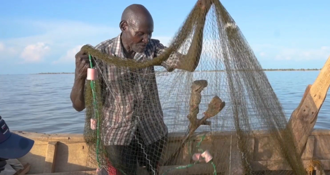 A man in a fishing boat lifts a fishing net, with crutches by his side
