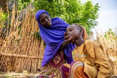 A woman, wearing a blue headscarf, bends over to wash the face of a boy sufering from an NTD .
