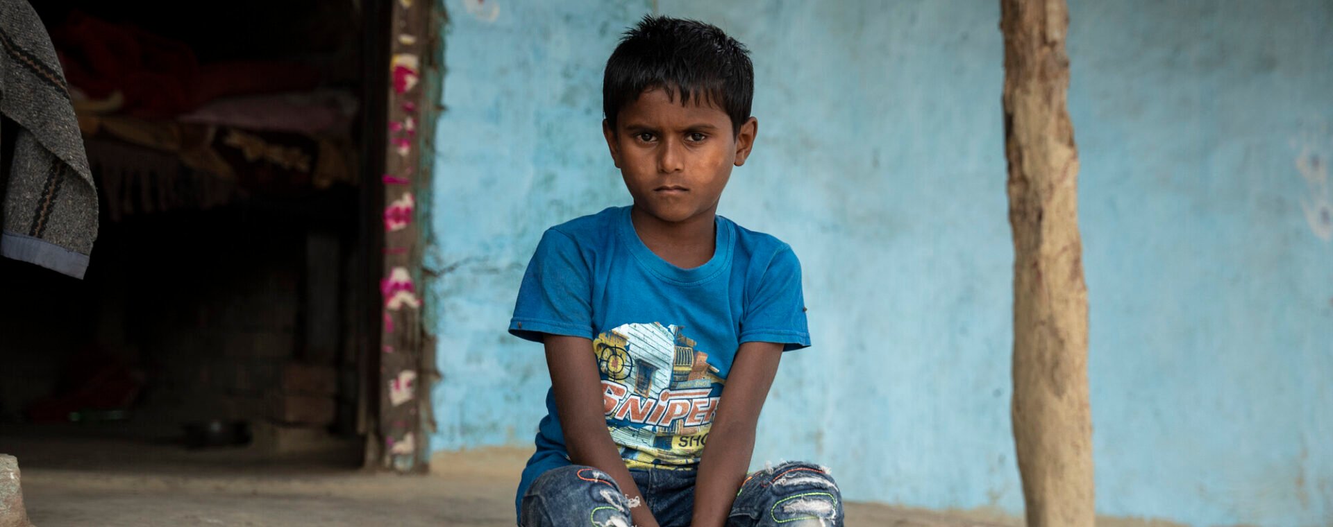 A little boy wearing a t-shirt and jeans sits on the porch of a blue painted house.